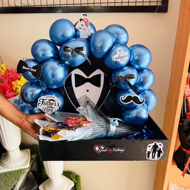 The Gentleman Dad Hamper featuring blue chrome balloons, assorted chocolate bouquet, tuxedo-themed backdrop and Fathers Day decorations in a premium black tray by Send My Feelings.
